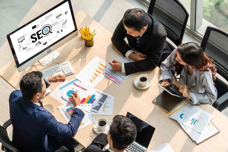 Grupo de personas en mesa de trabajo con tablets y documentos. En un computador se lee la palabra SEO