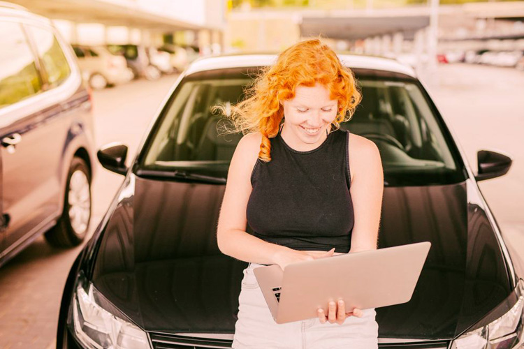 Mujer con un computador portátil sentada en el capót de un carro